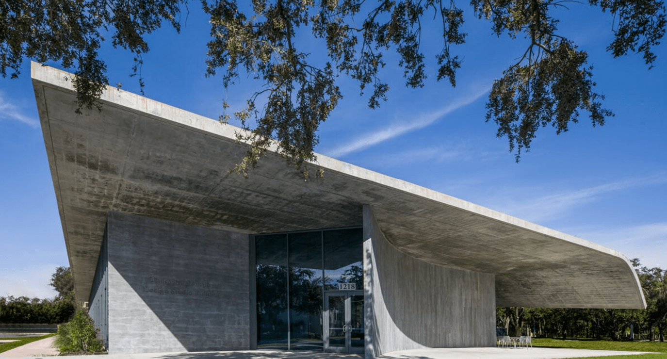 Modern concrete building with a sweeping roof, glass entrance, and trees framing a bright blue sky