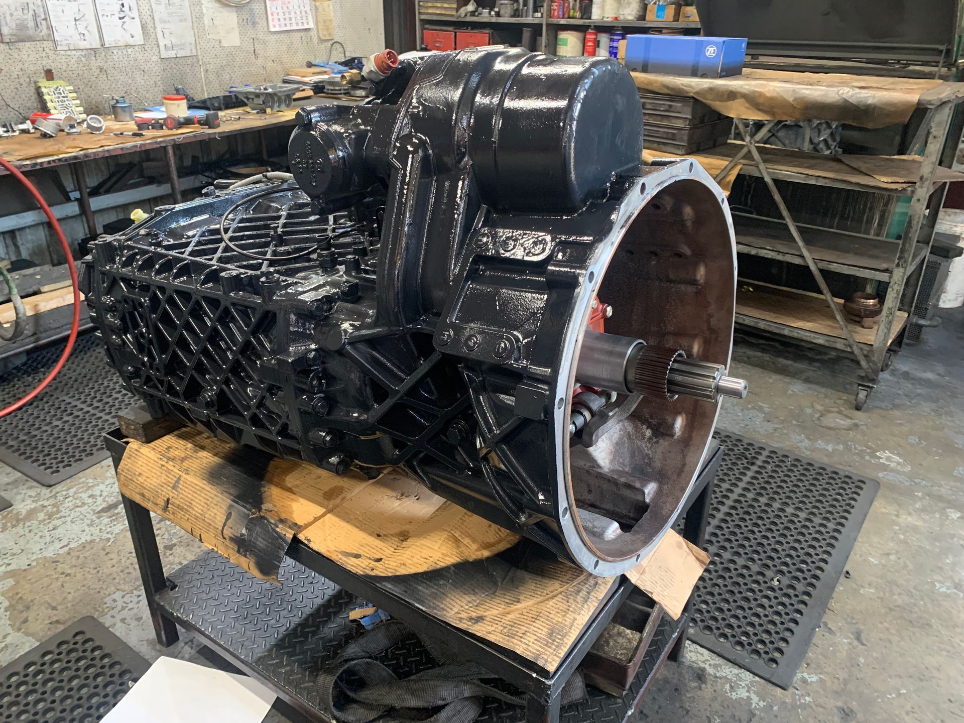 Black transmission on a workbench in a mechanic's shop, various tools and parts visible in the background.