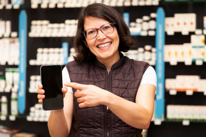 Eine Frau hält in einer Apotheke ein Mobiltelefon und zeigt darauf.