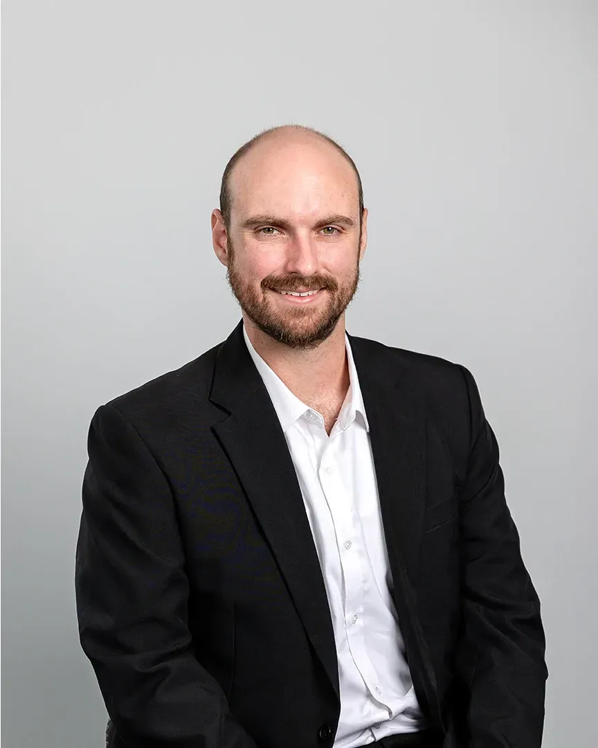 Studio portrait of a man in a black blazer and white shirt, seated against a light gray background