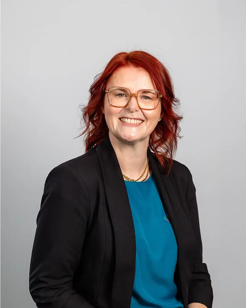 Portrait of a smiling person with red hair and glasses, wearing a black blazer over a teal top.