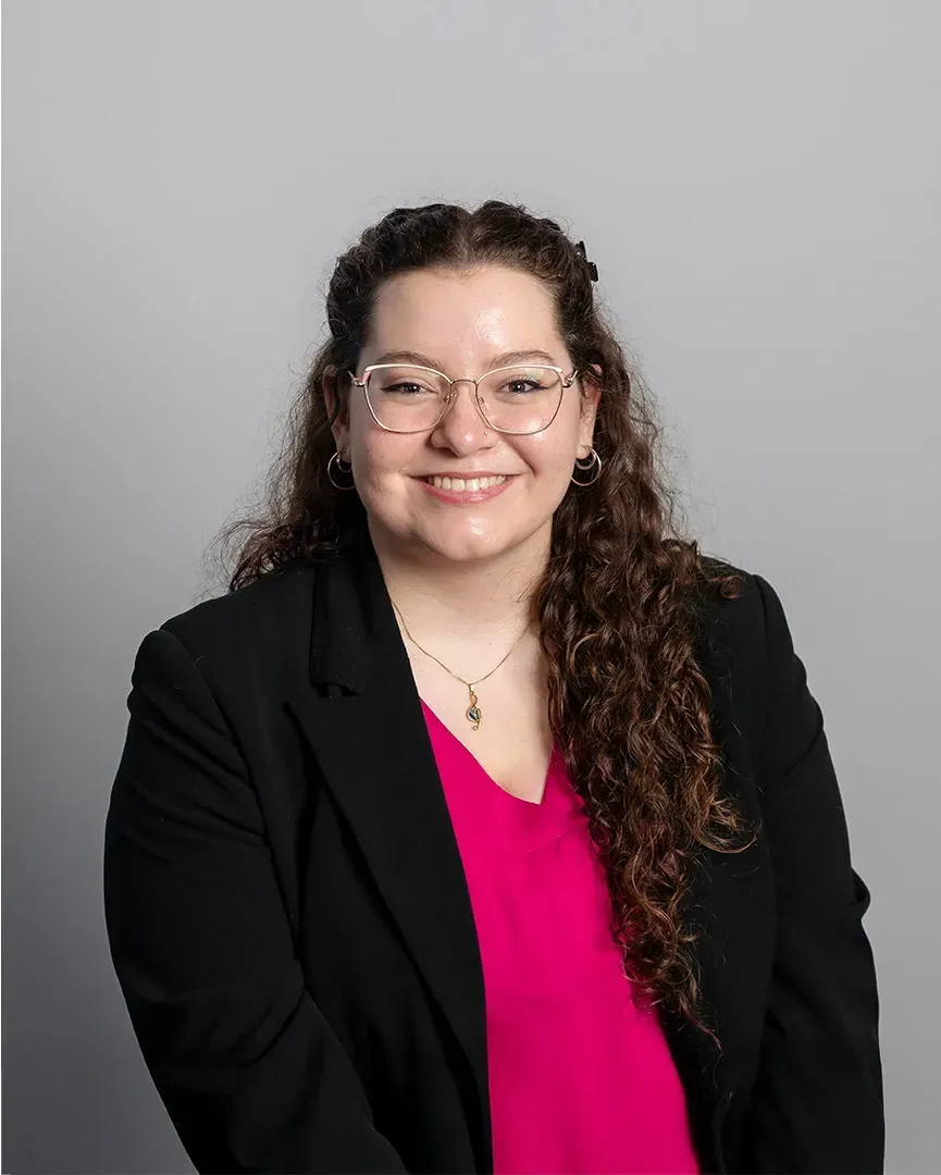 Woman in a black blazer and pink blouse smiling against a gray studio backdrop