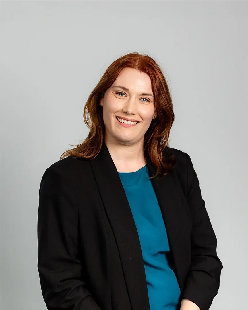 Smiling woman in a black blazer over a teal top against a light gray background