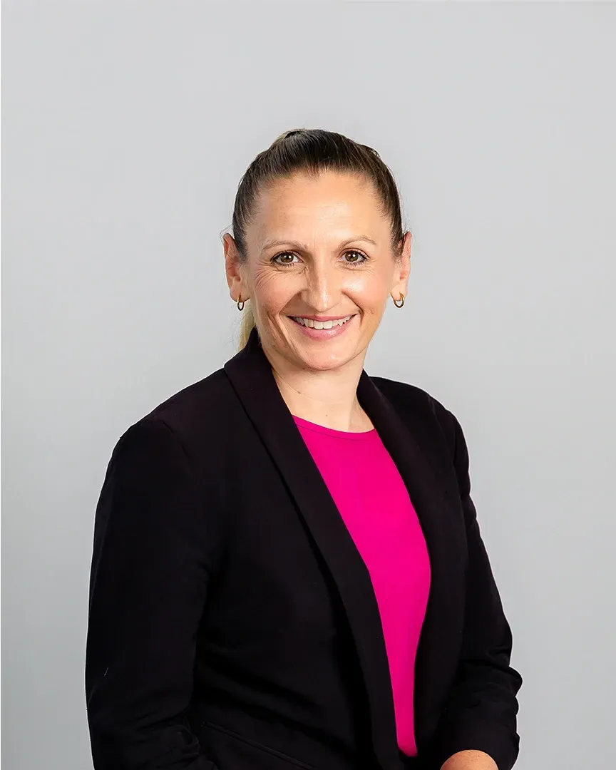 Smiling person in a black blazer and bright pink top against a light gray background