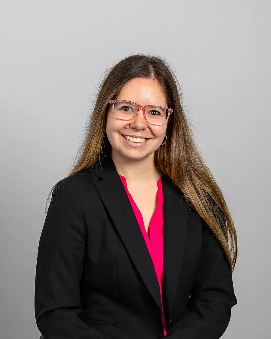 Smiling person in a black blazer and pink top, standing against a light gray background.