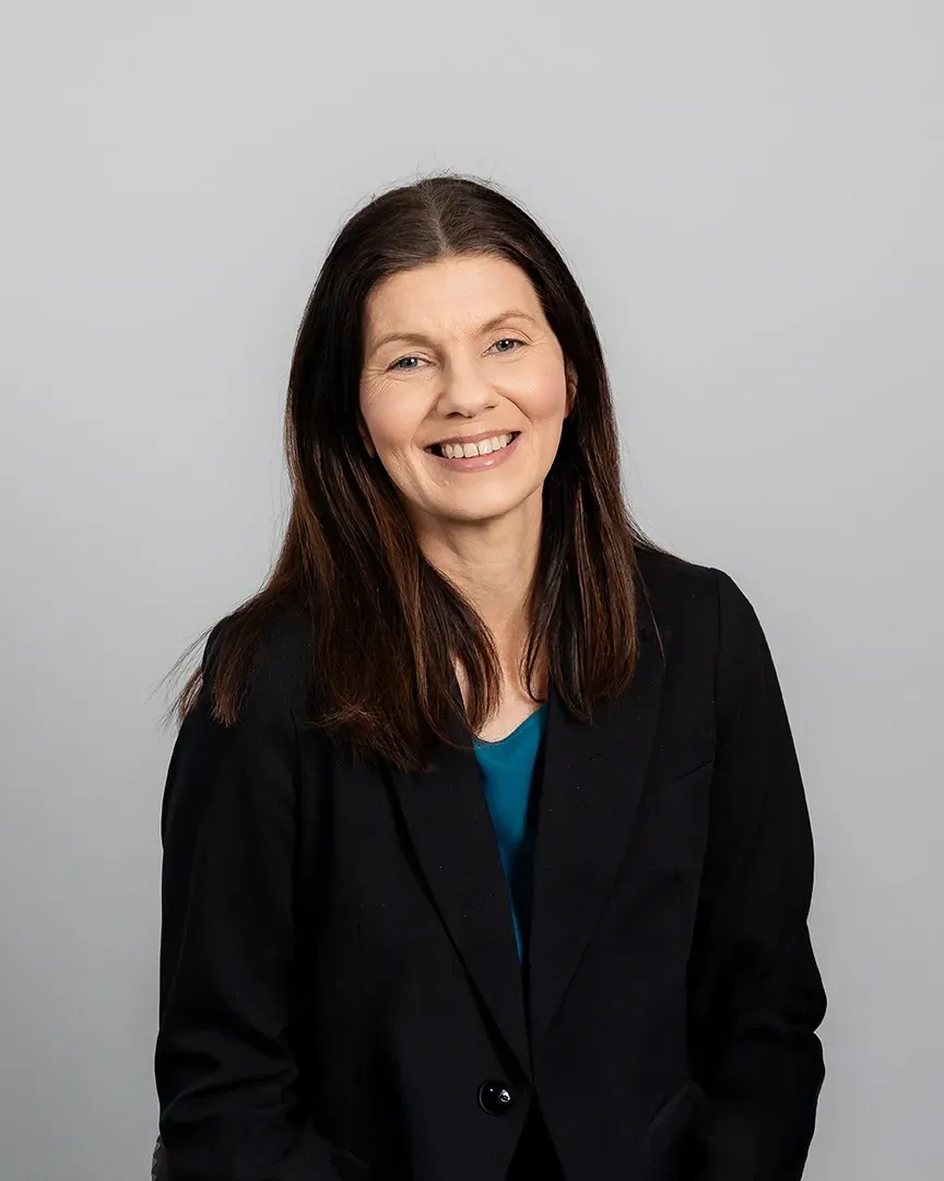 Portrait of a smiling person in a black blazer over a teal top against a gray background