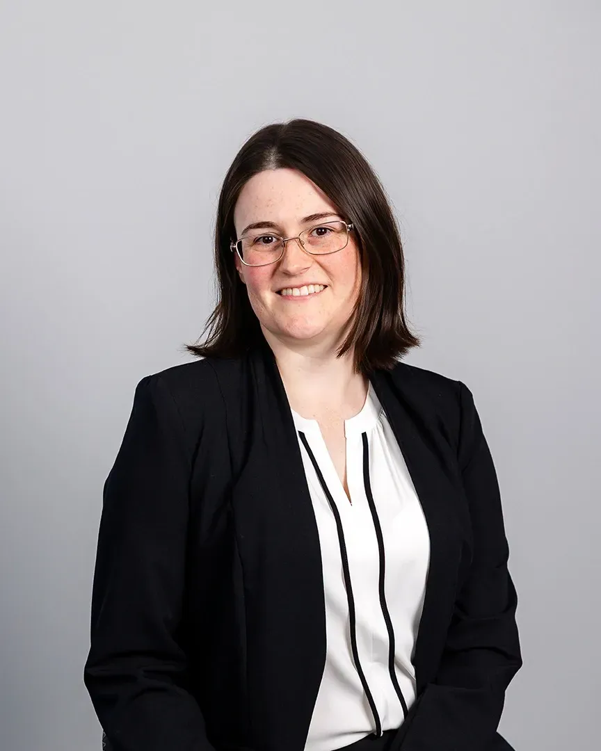 Portrait of a smiling person in a black blazer and white blouse against a gray background