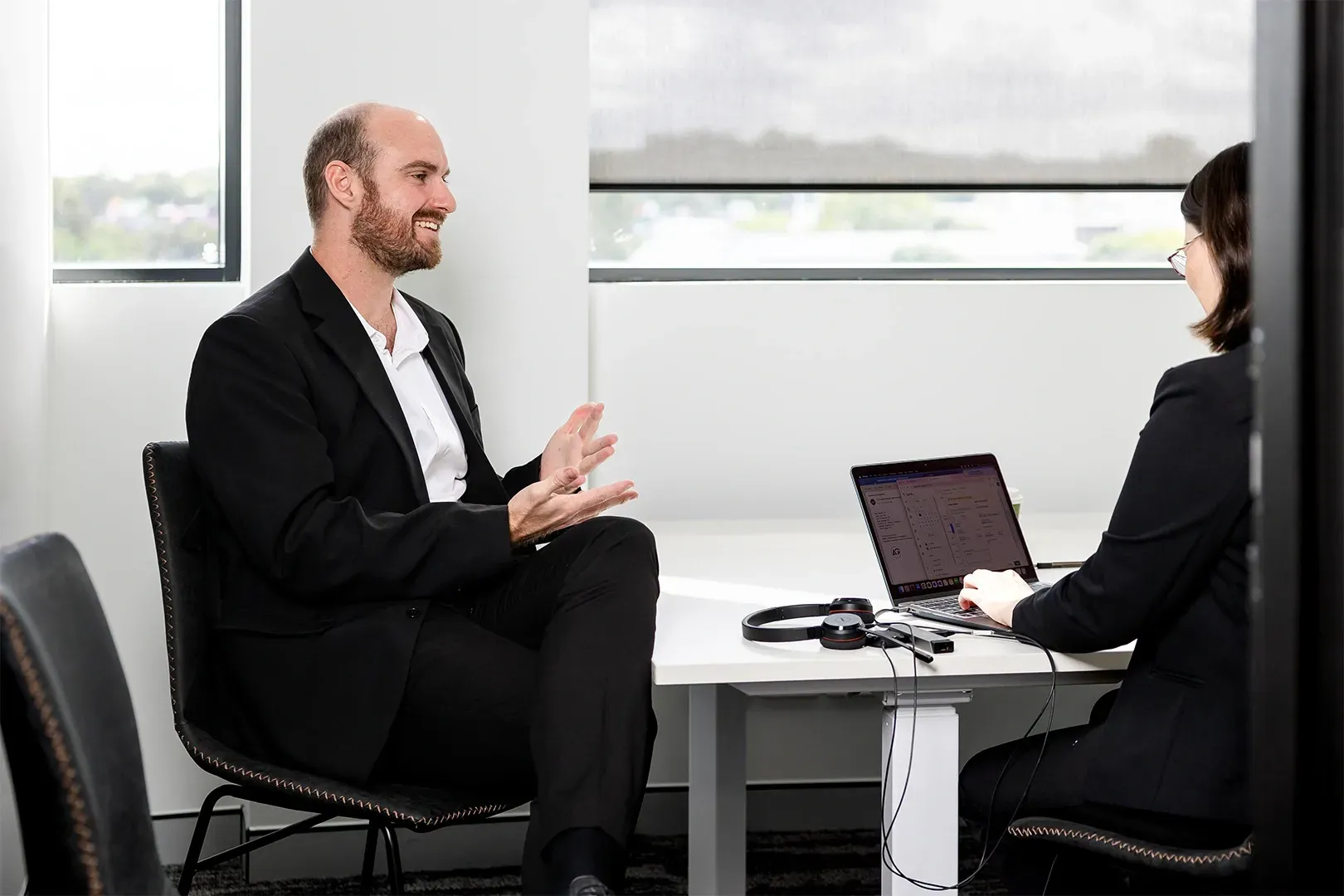 Two people in business attire meeting across a desk with a laptop in a bright office