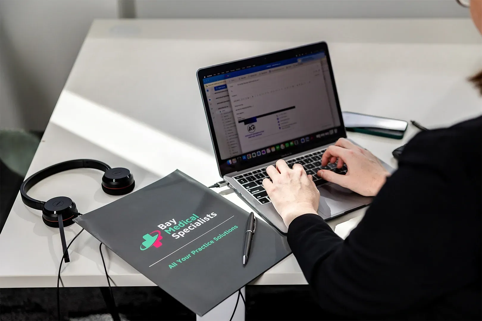 Person typing on a laptop beside headphones and a black folder on a white desk