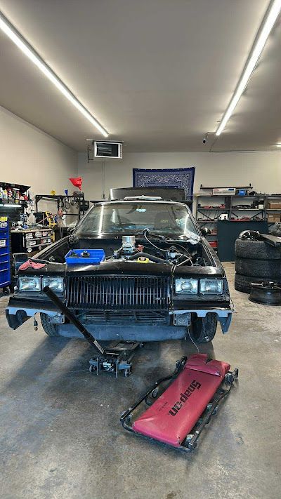 Black car being worked on in a garage; front end disassembled, supported by a jack. 