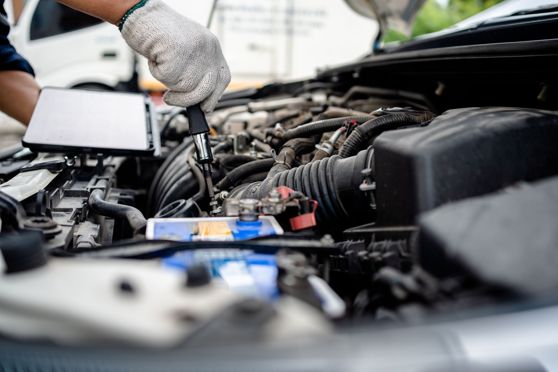 Mechanic working on a car engine, holding a tool and tablet, outdoors, sunny day.