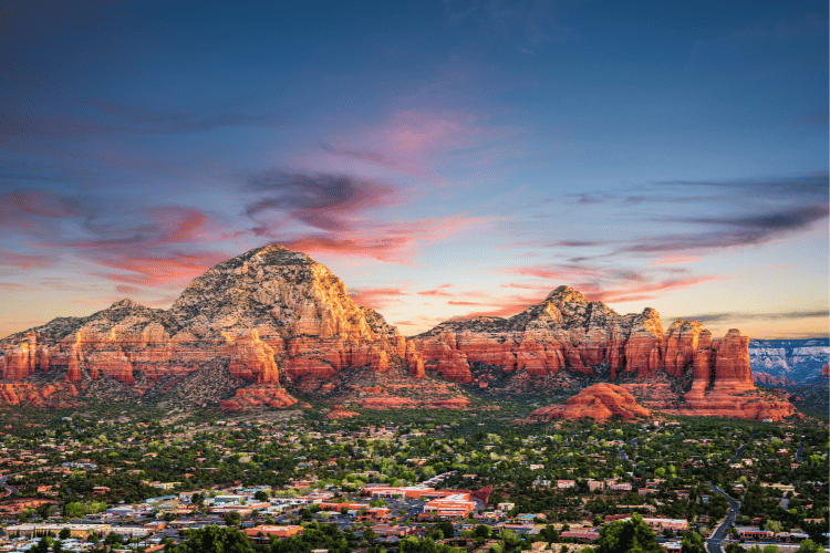 Sedona, Arizona landscape with rugged red rock formations illuminated by a soft, colorful sunset above a forested town.