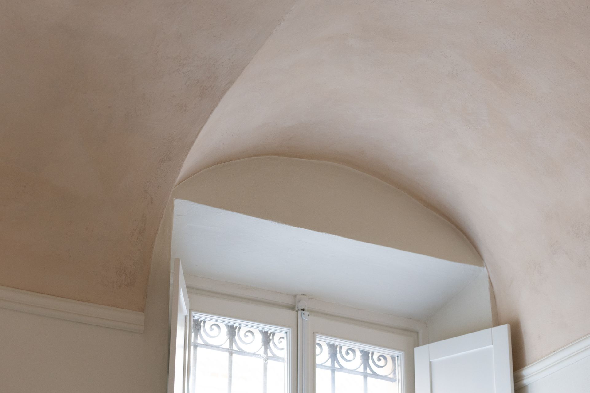 Soffitto a volta sopra una finestra bianca con inferriate metalliche decorate e persiane aperte.