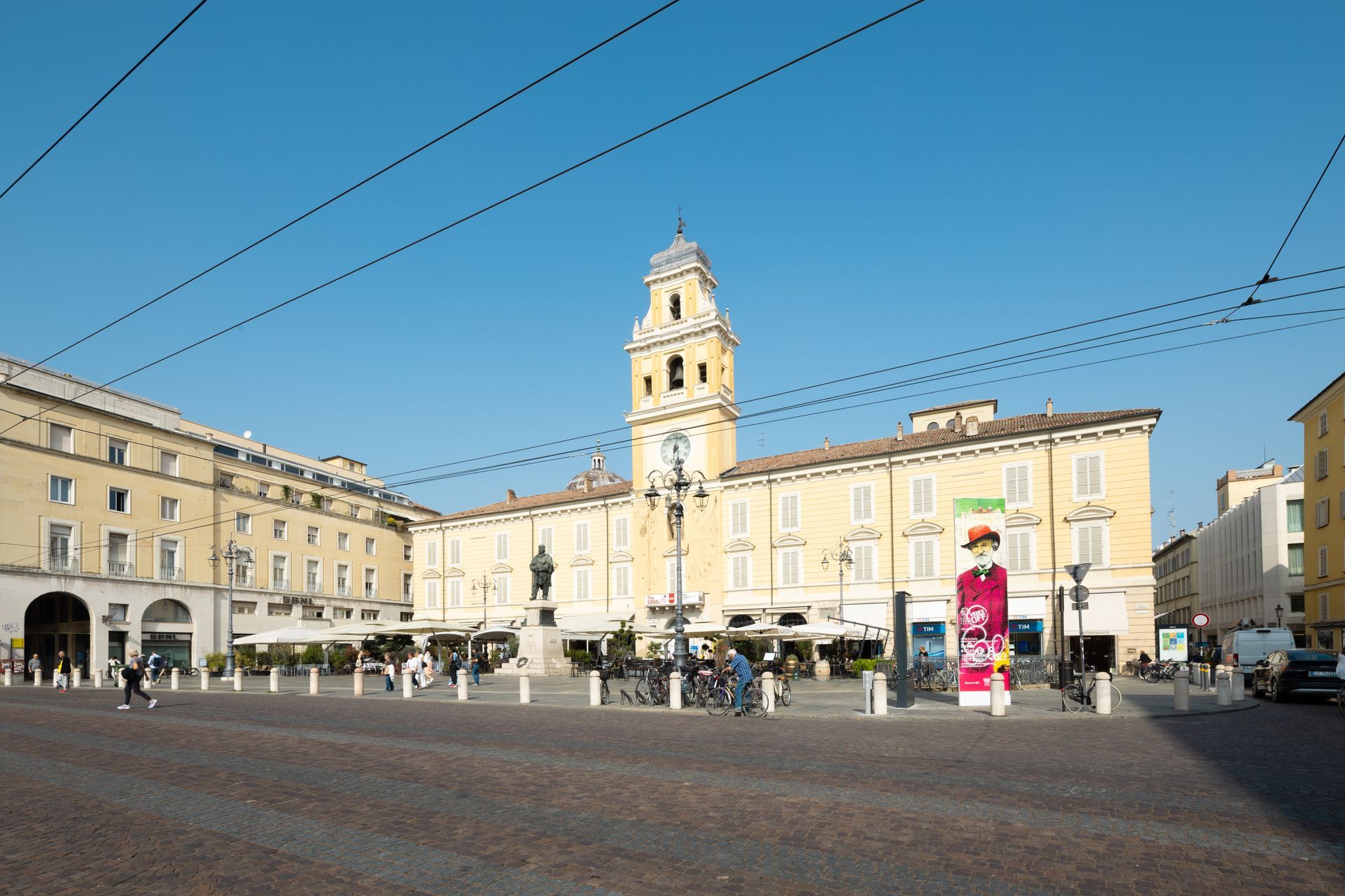Piazza europea soleggiata con edifici chiari, una torre dell'orologio e cavi del tram aerei.