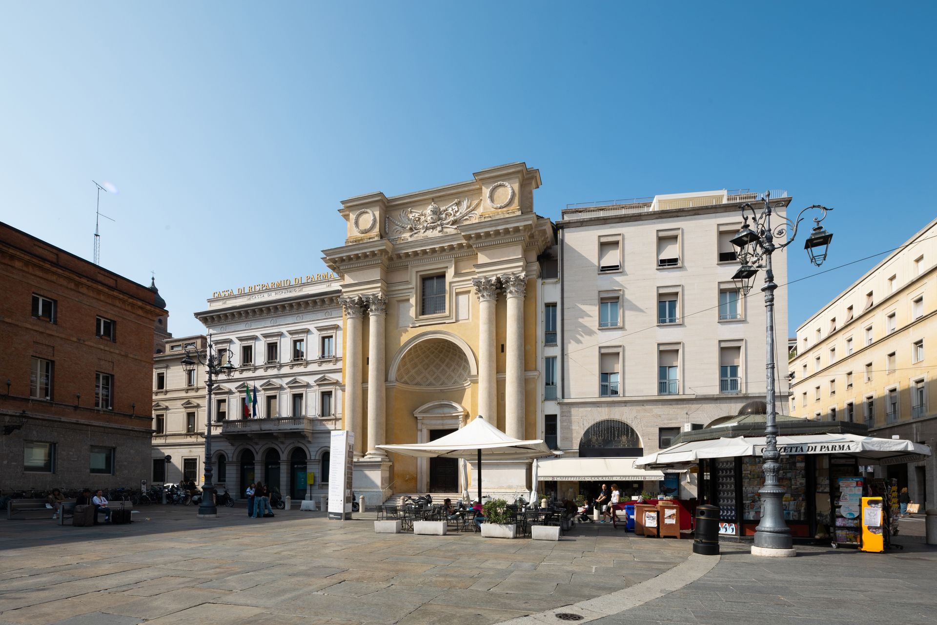 Piazza storica con un grande arco decorato, edifici circostanti e tavolini all'aperto sotto un cielo azzurro e limpido.