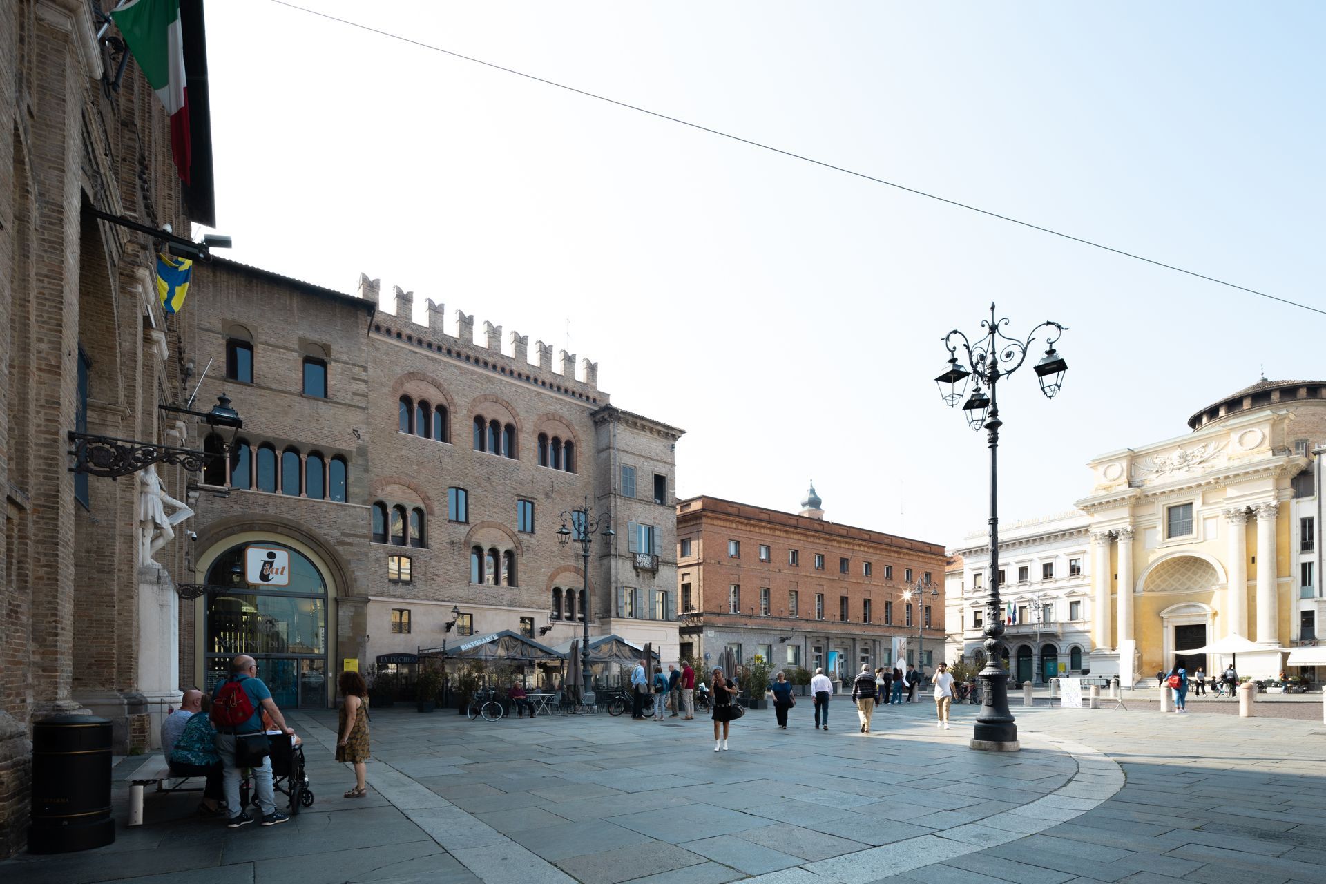 Piazza europea soleggiata con edifici storici, un lampione centrale e alcuni pedoni che la attraversano.
