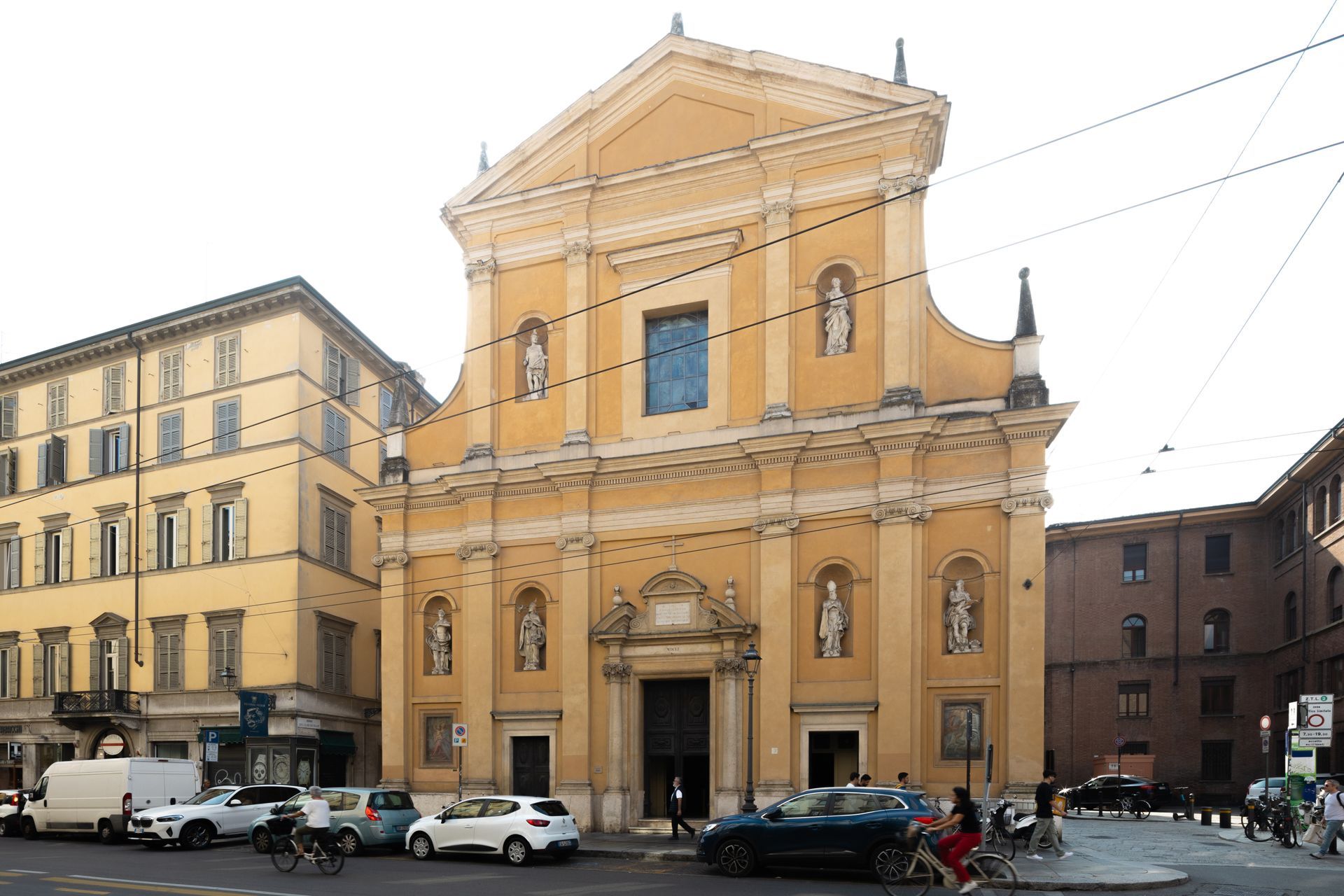 Chiesa storica gialla in una piazza cittadina, con auto parcheggiate ed edifici circostanti sotto un cielo luminoso