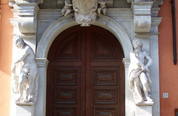 Un'elegante doppia porta in legno, incorniciata da un arco in pietra e da statue, si erge sulla facciata di un edificio color pesca.