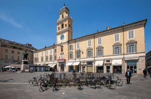 Piazza cittadina soleggiata con un edificio storico beige e una torre dell'orologio, biciclette parcheggiate davanti e alcuni pedoni.