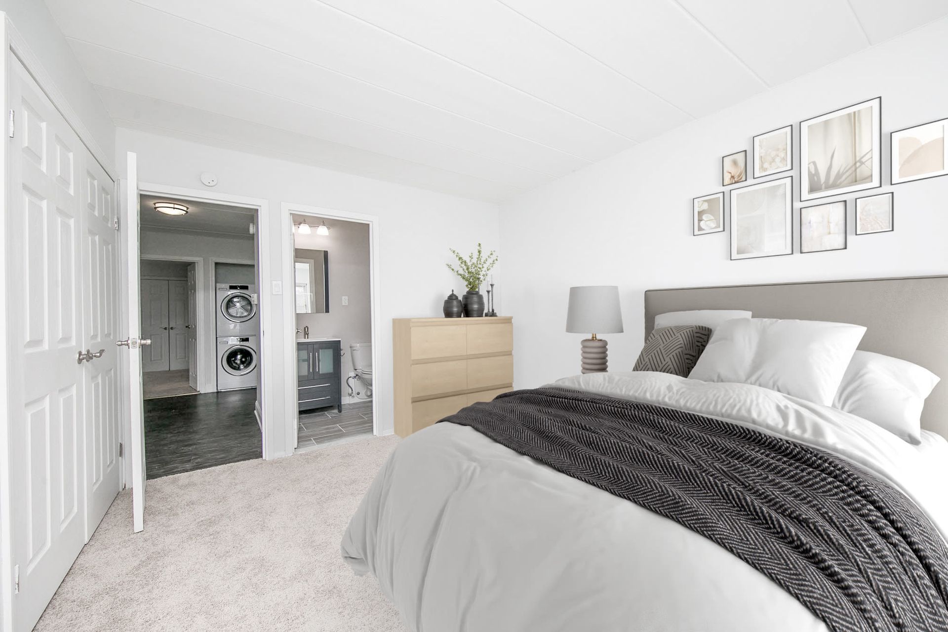 Bright bedroom with a large bed, dresser, lamp, and an open doorway to a bathroom and laundry area.