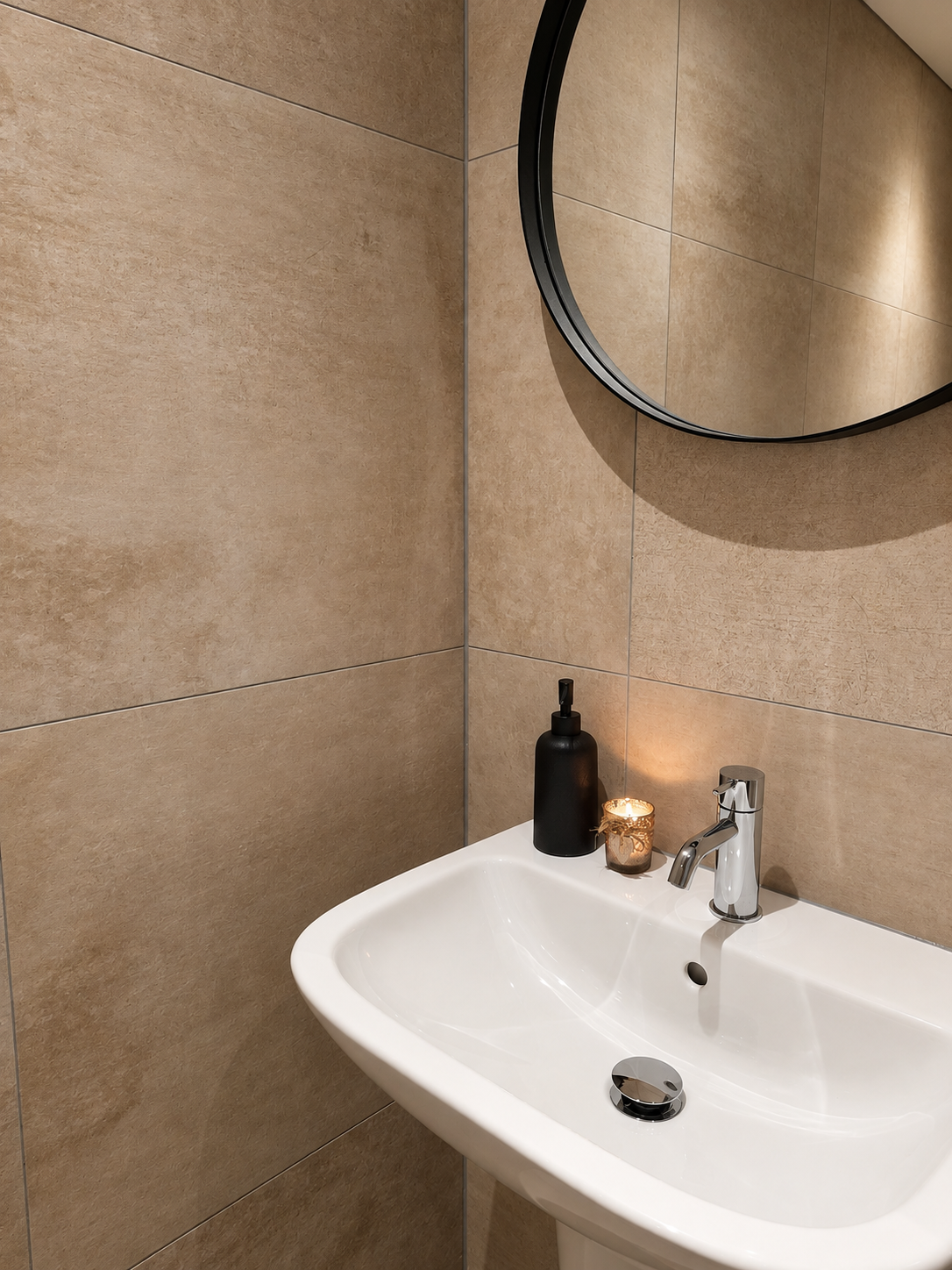 Moderne badkamer met wastafel, beige tegelwanden, ronde spiegel en zwarte zeepdispenser.