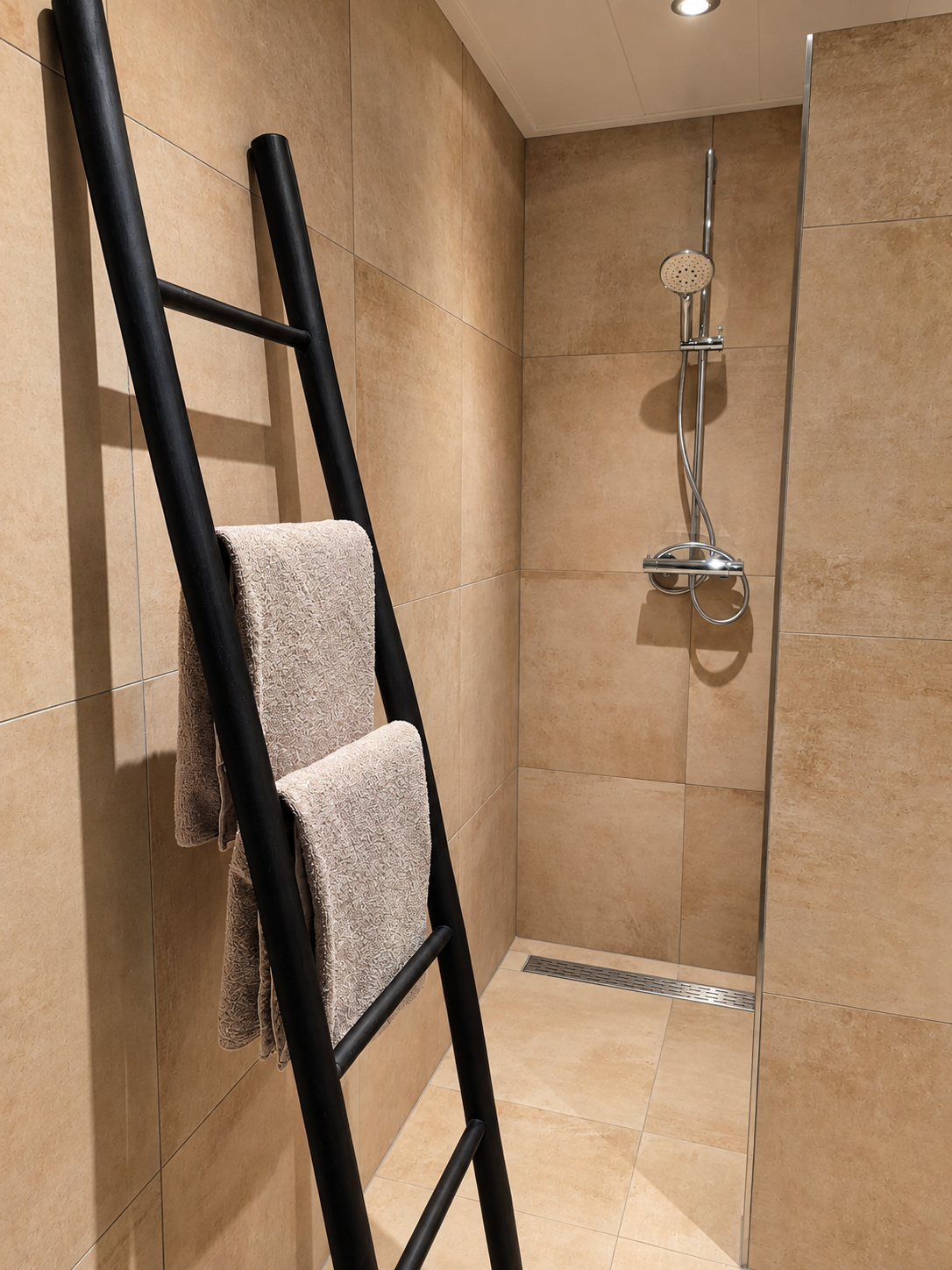 Moderne beige betegelde douche met een zwart handdoekrek in ladderstijl en een chromen douchekop.