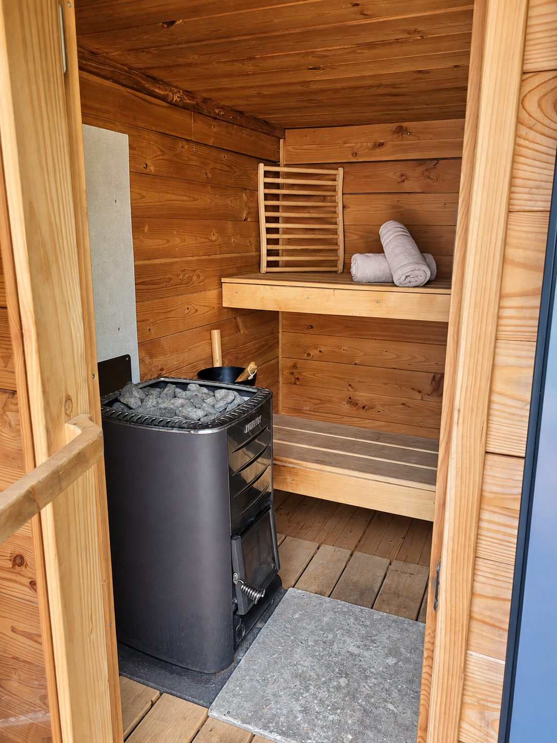 Houten sauna-interieur met kachel, saunastenen, bankje en handdoeken op een plank.