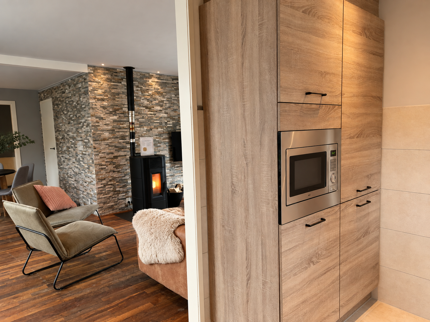 Moderne woonkamer met stenen open haard, fauteuil en houten lambrisering in de keuken met ingebouwde oven.