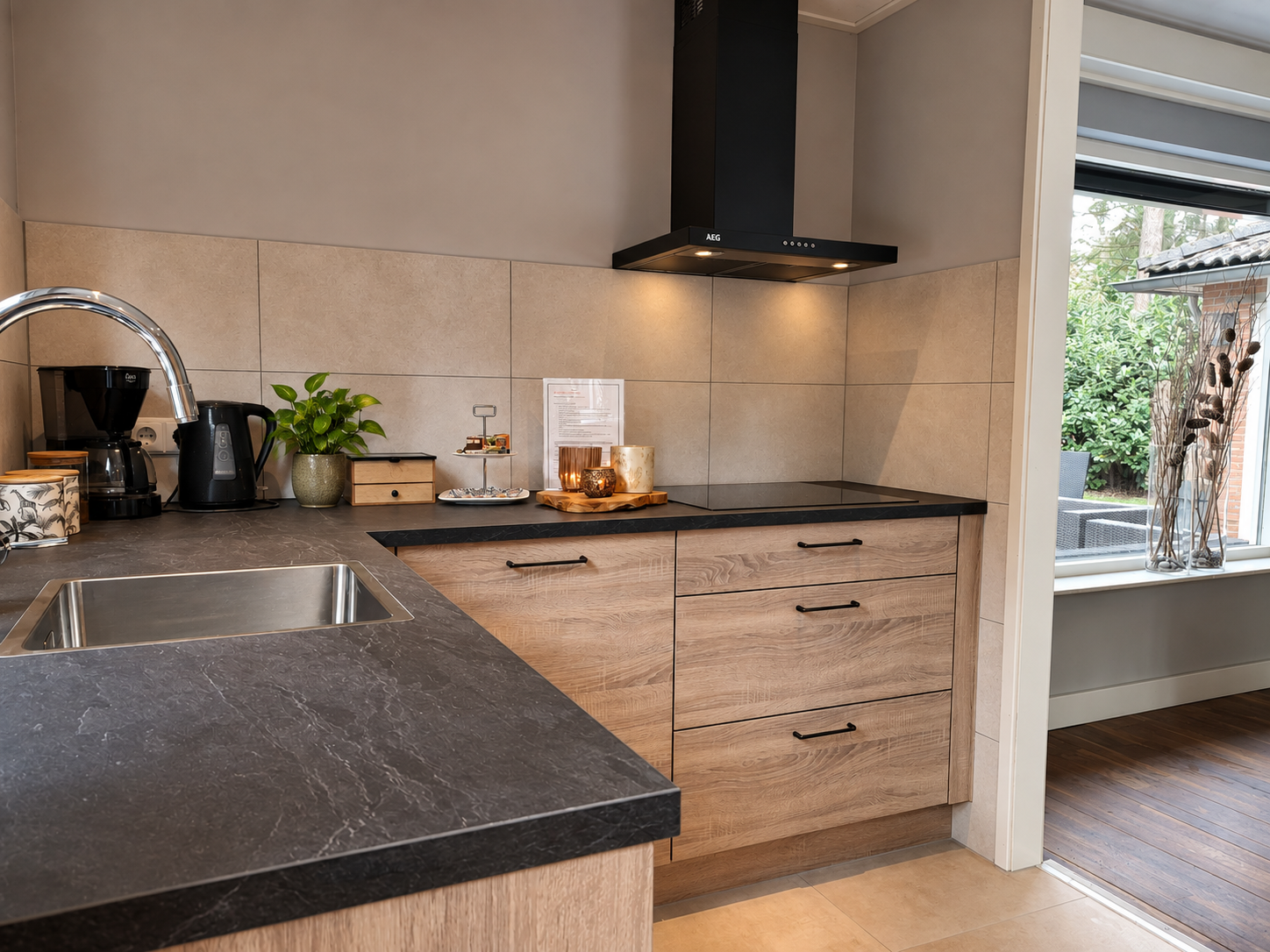 Moderne keuken met donkere werkbladen, lichte houten kasten, zwarte afzuigkap en een zitgedeelte bij het raam.