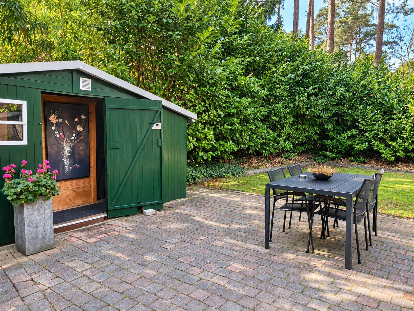 Groene tuinschuur met open deur naast een terrastafel en stoelen op een betegelde binnenplaats.