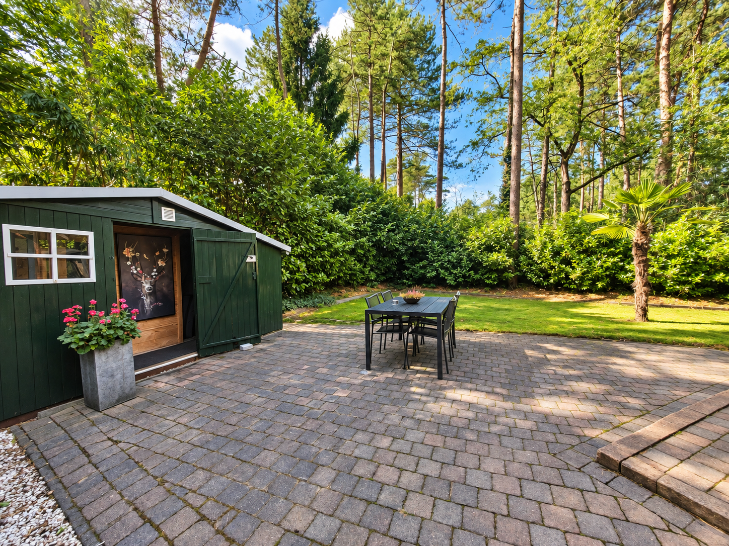Terras in de achtertuin met bestrating, een klein tafeltje en een groen tuinhuisje naast bomen en gazon.