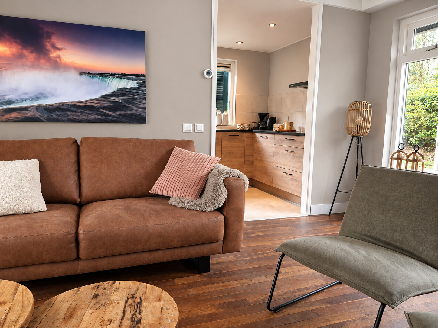 Gezellige woonkamer met bruine bank, grijze fauteuil, houten vloer, muurdecoratie met oceaanmotief en open doorgang naar de keuken.