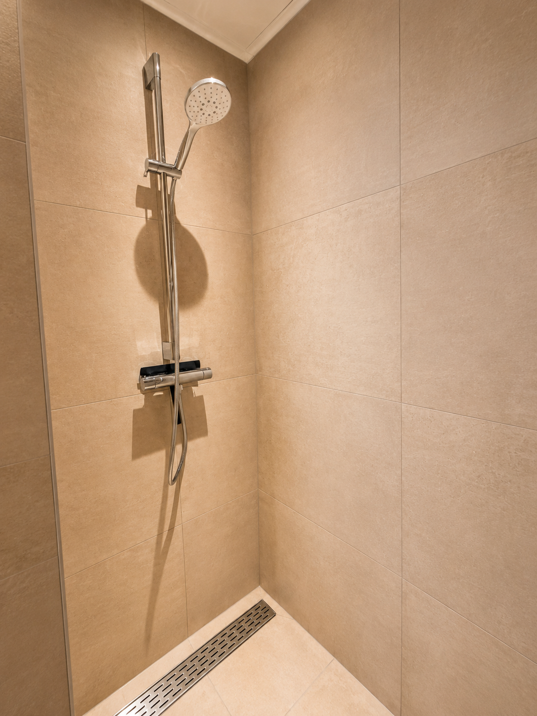 Inloopdouche met handdouche, ingebouwde mengkraan en lineaire vloerafvoer in beige tegels.