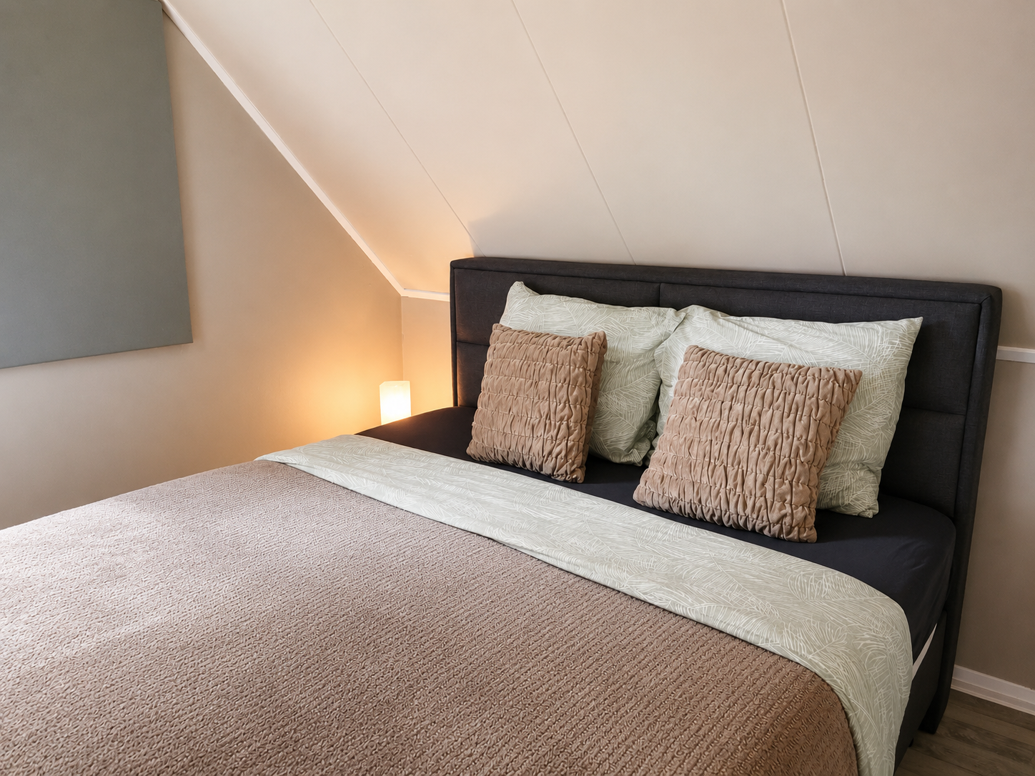 Een keurig opgemaakt bed in een knusse zolderkamer met beige beddengoed en een donker gestoffeerd hoofdeinde.