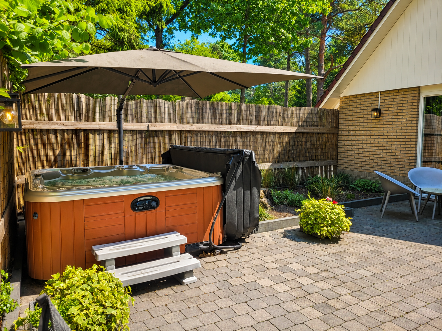 Jacuzzi met parasol op een betegeld terras in de achtertuin, naast een huis met een houten schutting.