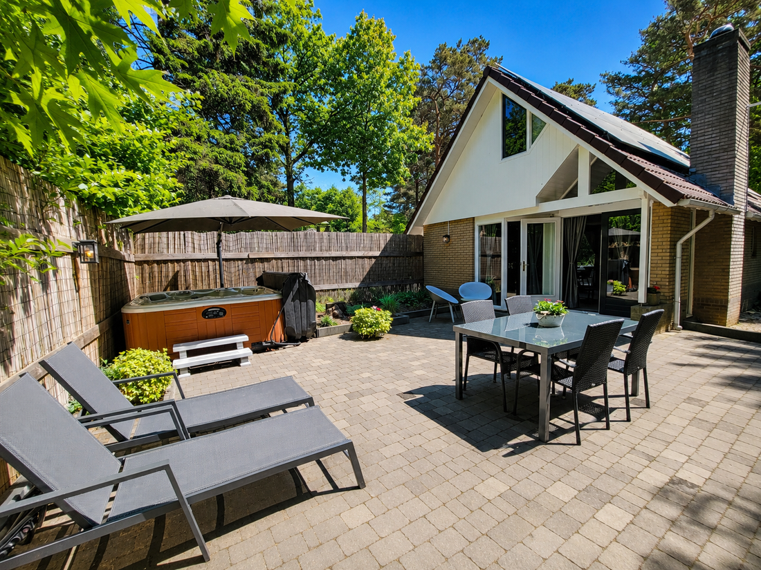 Zonnig terras in de achtertuin met ligstoelen, eettafel, jacuzzi en een klein huisje met openslaande deuren.