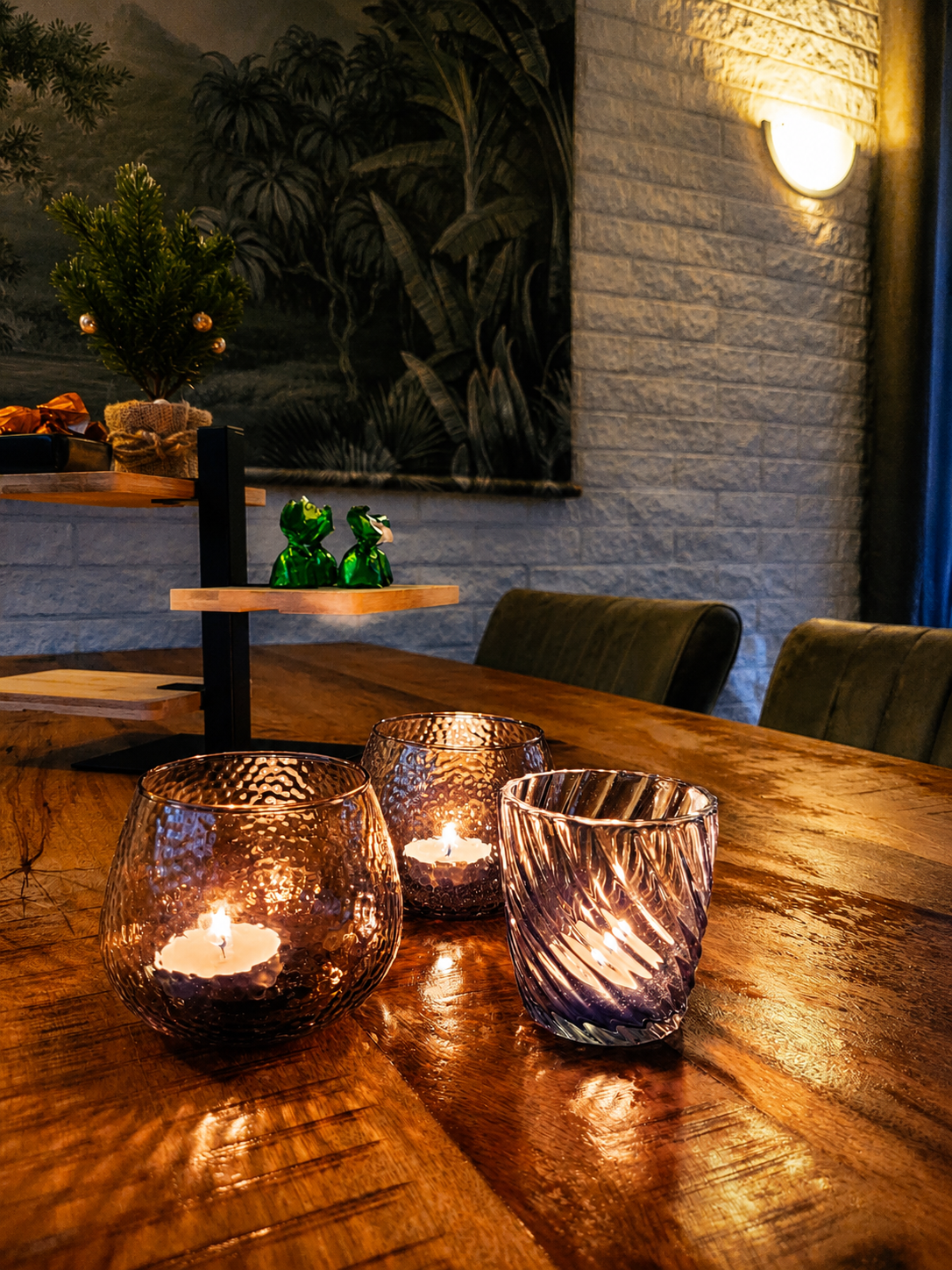 Een houten tafel bij kaarslicht op een terras met drie glazen kaarsenhouders en een klein groen kikkerbeeldje.