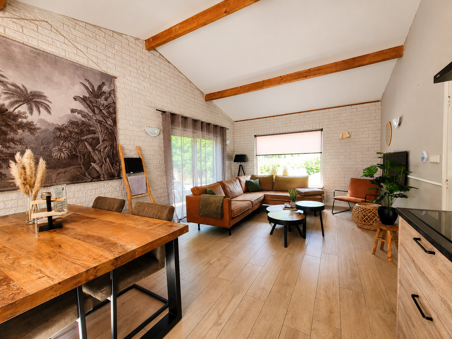Moderne woonkamer met houten balken, witte muren, beige bank, houten bureau en grote ramen.