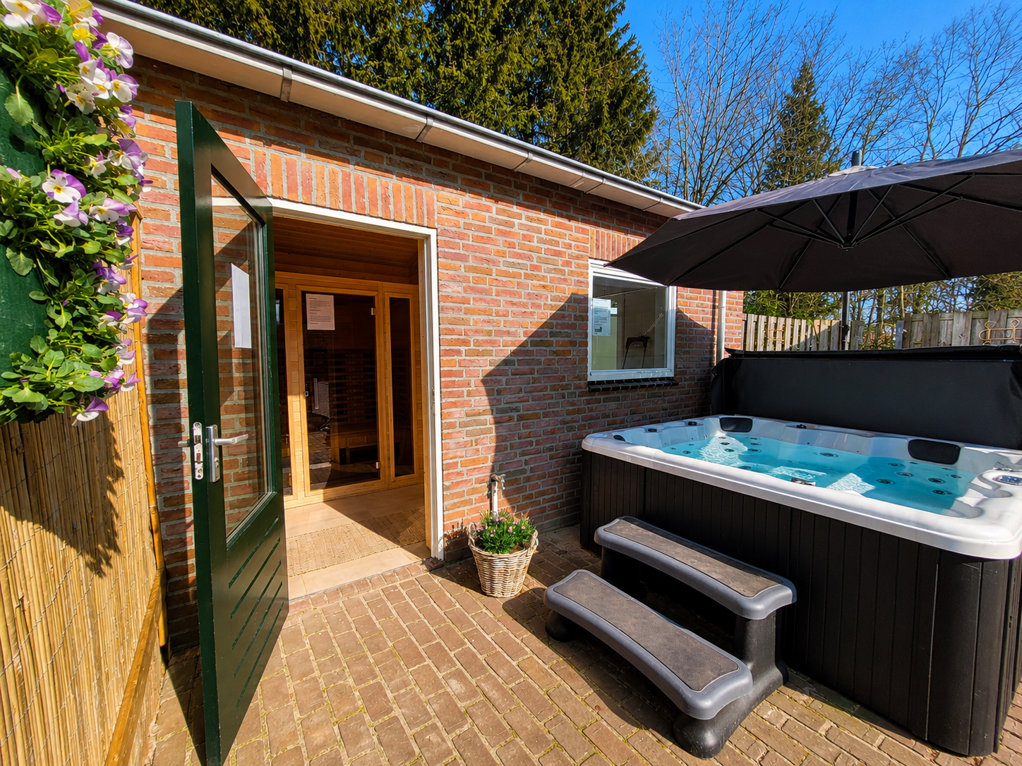 Terras in de achtertuin met jacuzzi, stenen bijgebouw, openslaande deur en trap onder een overkapping.