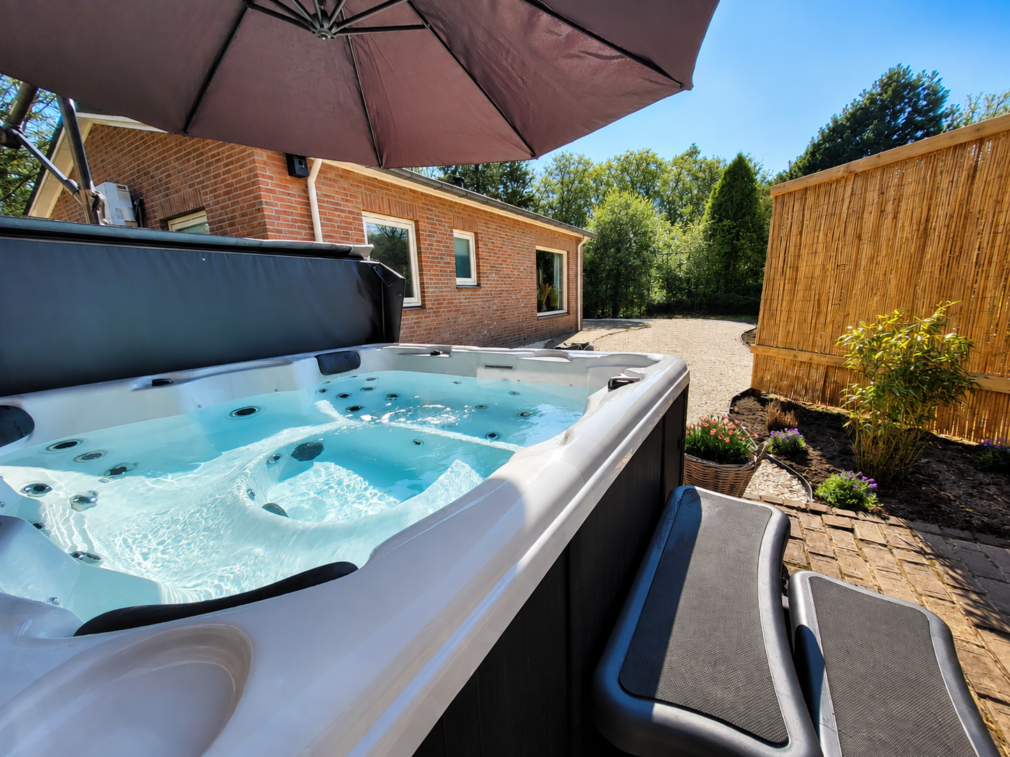 Een bubbelbad met bubbels in de achtertuin, onder een parasol, naast een bakstenen huis met een houten schutting.