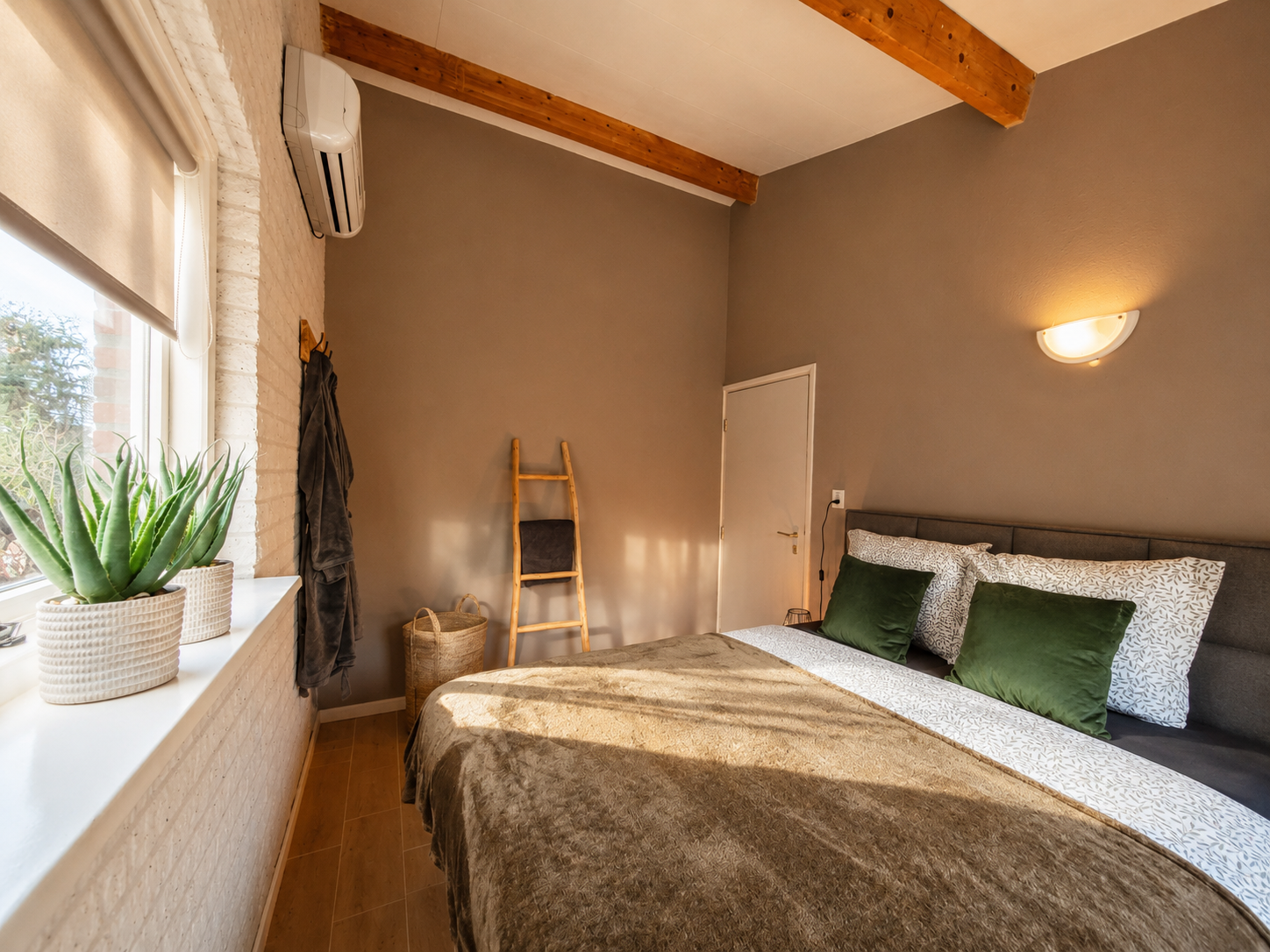 Gezellige slaapkamer met beige muren, een bed, wandlamp, houten balken en een ladder die tegen het raam leunt.