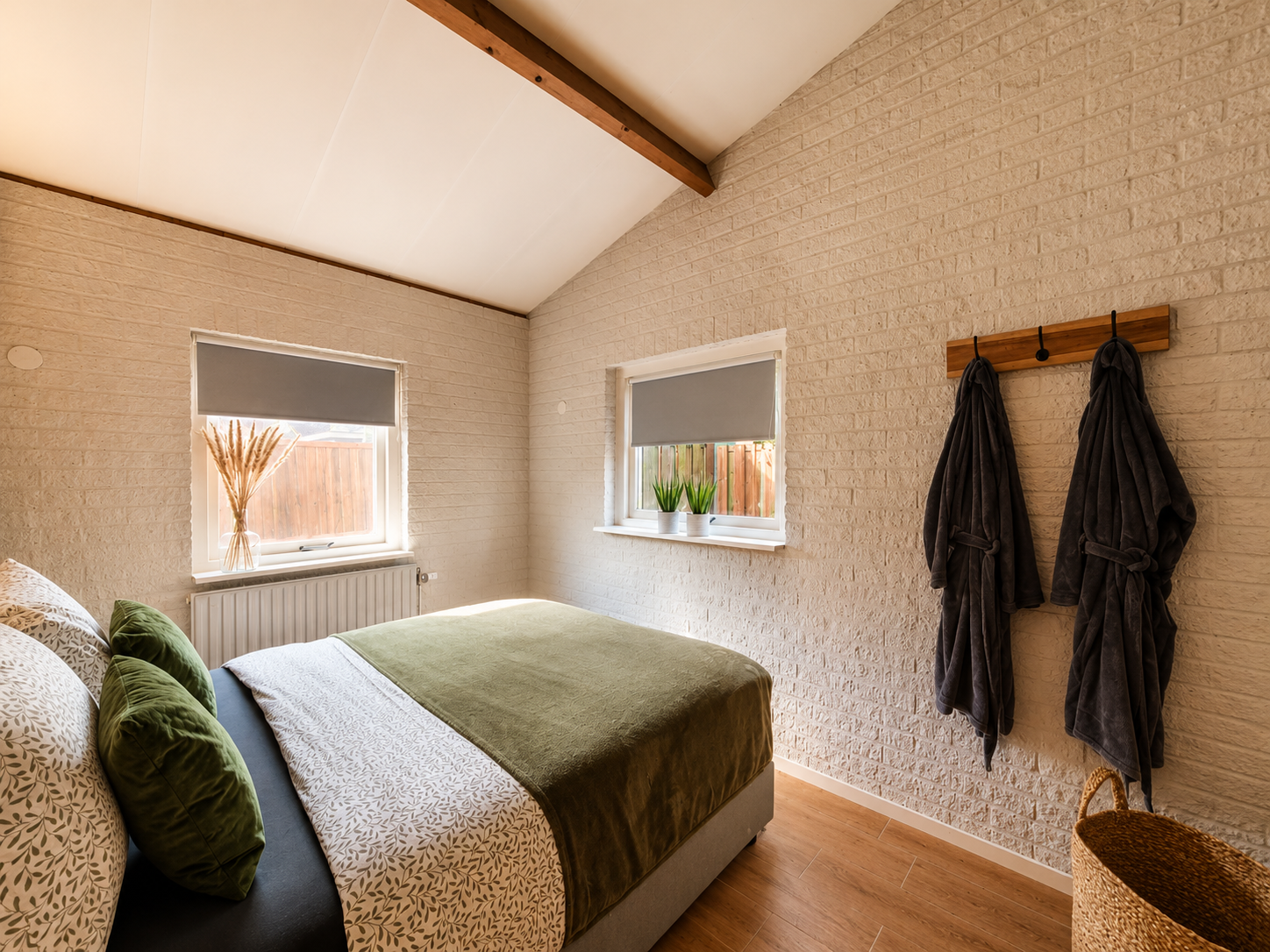Gezellige slaapkamer met groene sprei, twee kleine ramen en haken aan de muur waaraan donkere badjassen hangen.