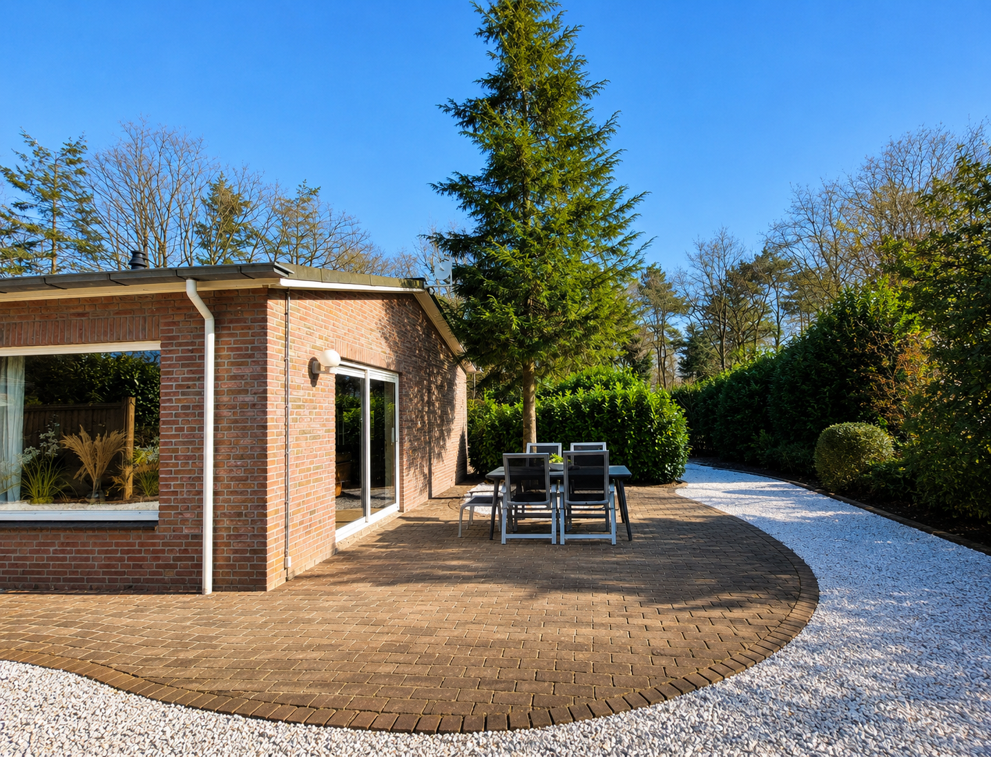 Een stenen terras met een eettafel, een grindpad en hoge groenblijvende bomen in een zonnige tuin.