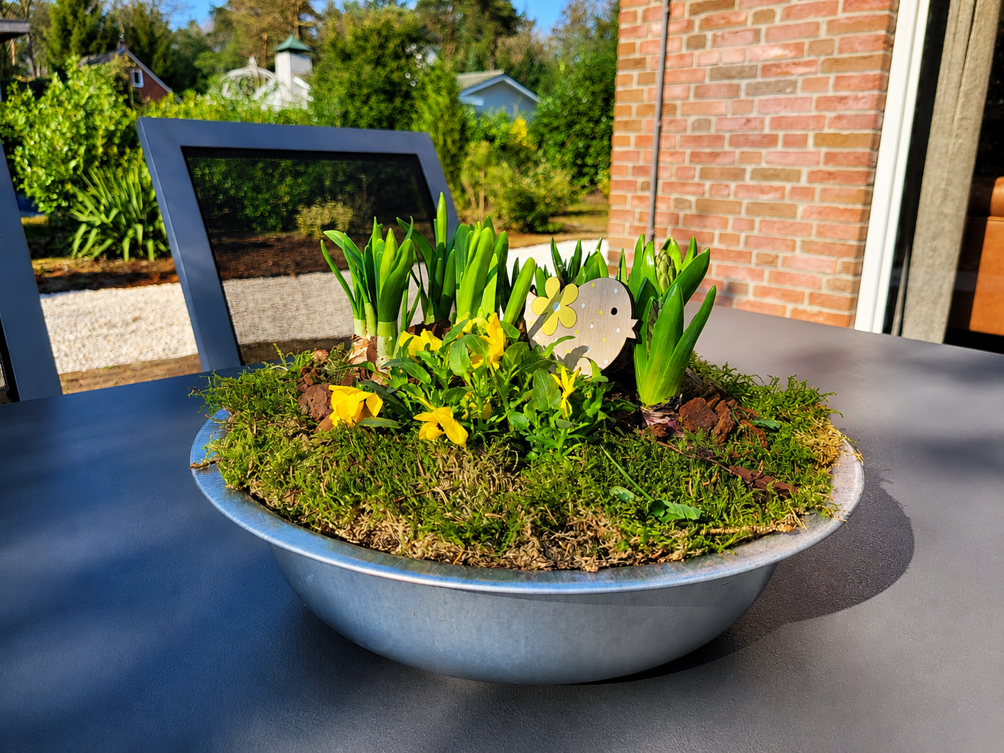 Een kleine plantenbak met groene planten en gele bloemen op een terras bij een bakstenen muur.