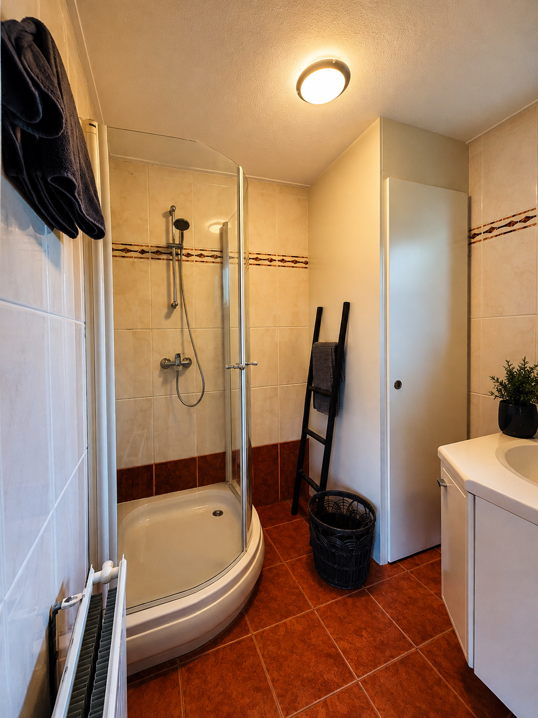 Kleine badkamer met douchebad, glazen scheidingswand, rode tegelvloer en wastafelmeubel.