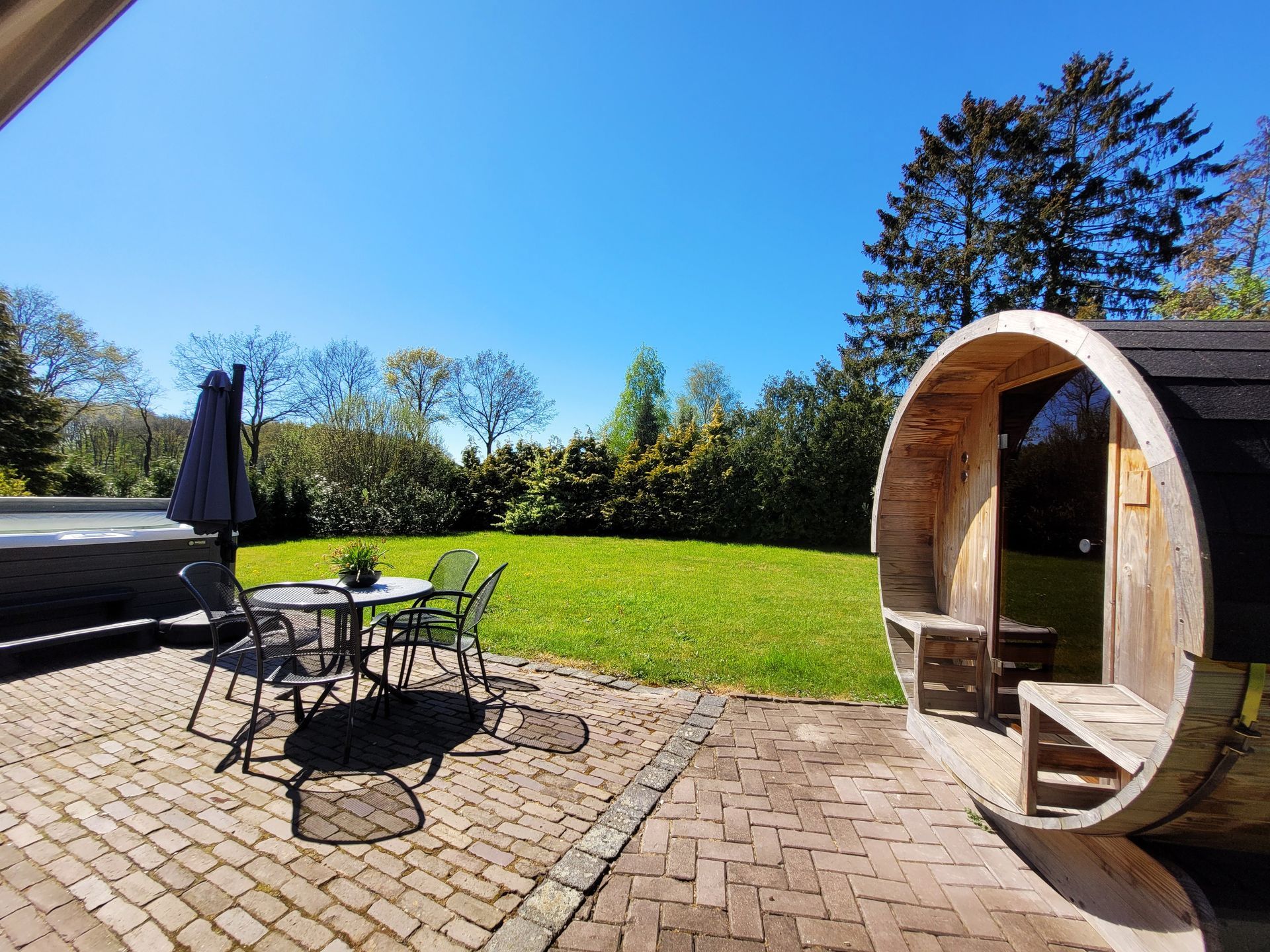 Terras met tafel, stoelen, parasol en houten saunaton naast een grasveld onder een blauwe hemel.