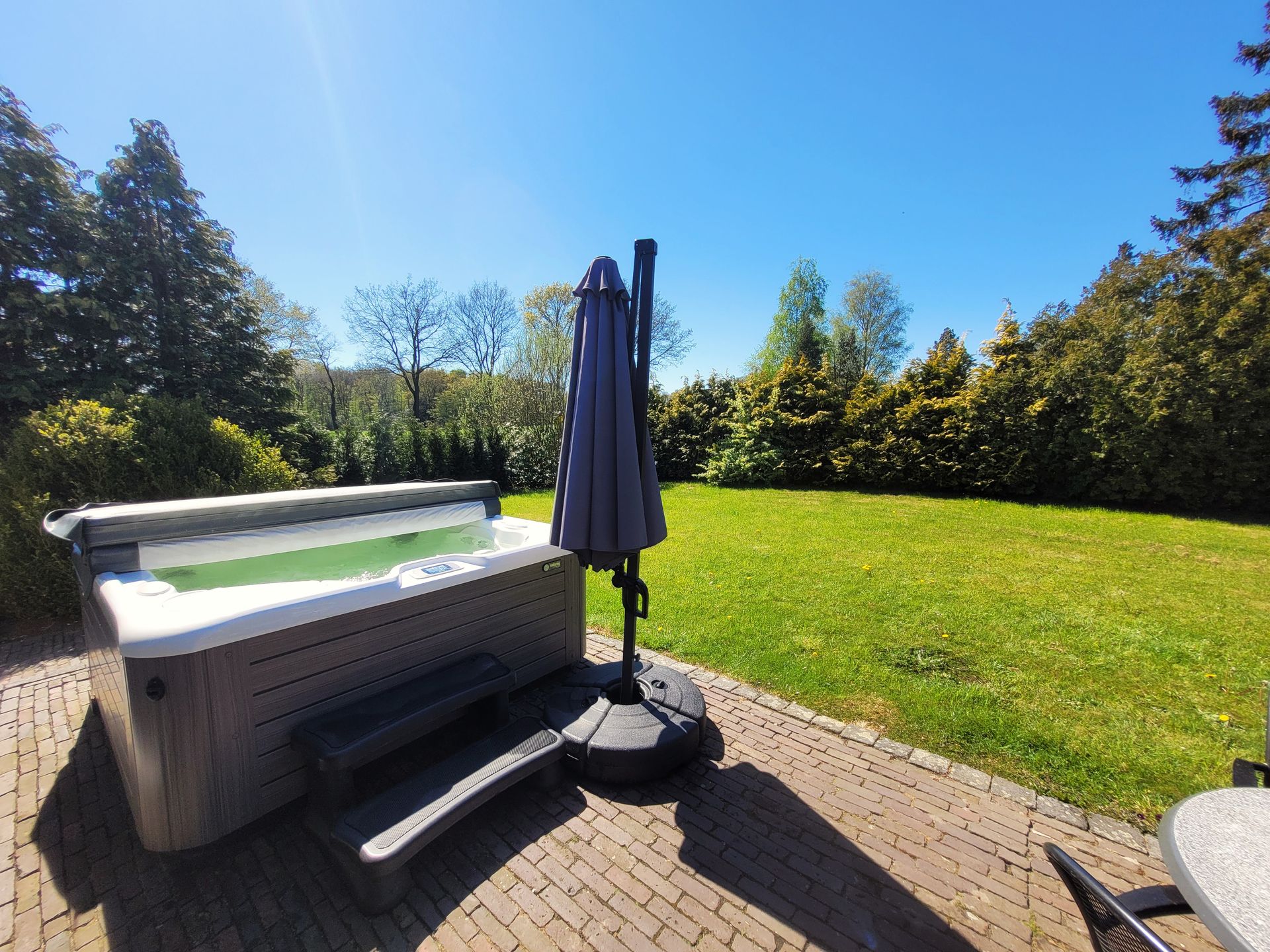 Terras in de achtertuin met een jacuzzi, een gesloten parasol en een groen gazon onder een stralend blauwe hemel.
