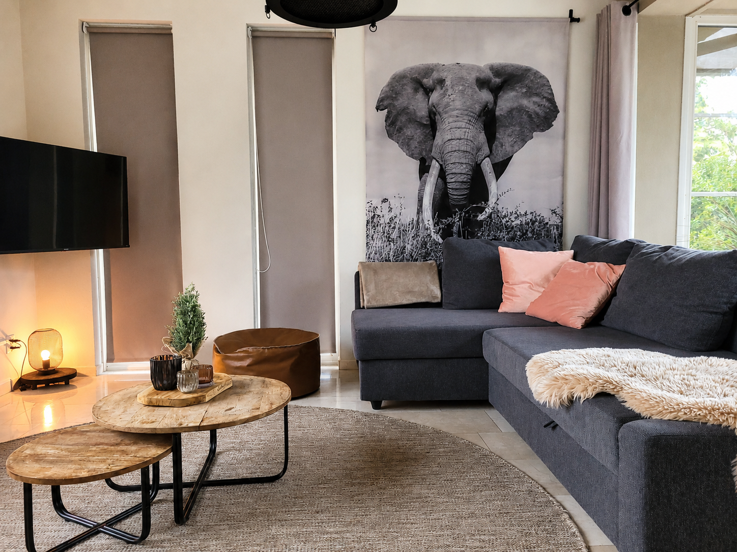 Gezellige woonkamer met grijze hoekbank, ronde salontafels, lamp en een groot olifantenkunstwerk aan de muur.