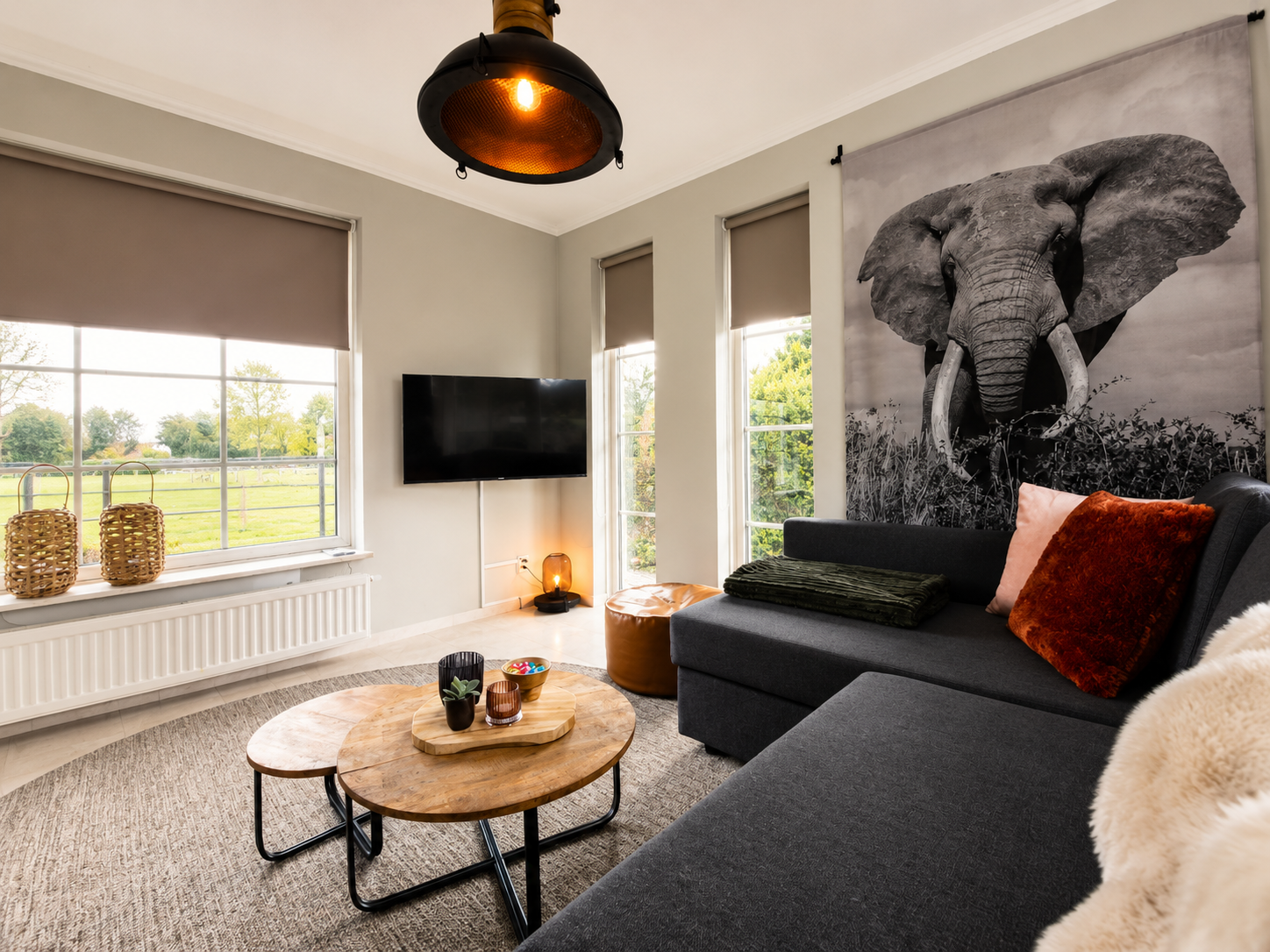 Lichte woonkamer met olifantenmuurdecoratie, grijze hoekbank, ronde salontafel en raamraam.