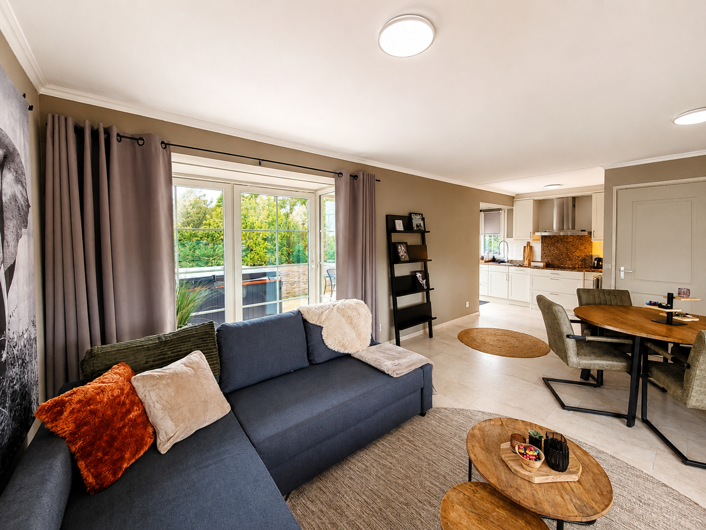 Lichte woonkamer met blauwe bank, groot raam, eettafel en open keuken.